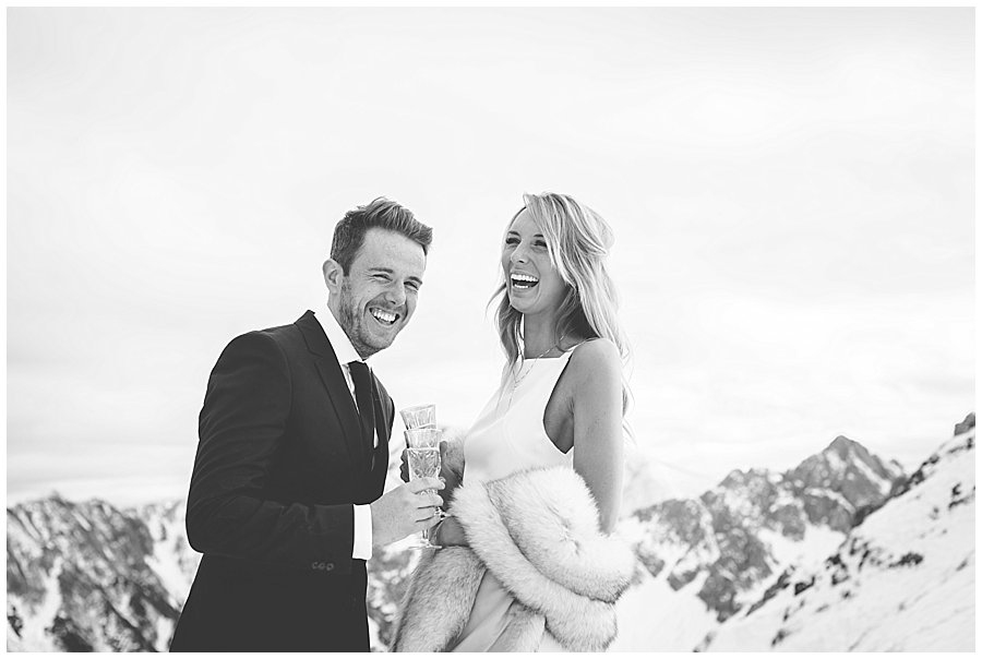 Steph and Lee start laughing holding champagne glasses with mountains in the background