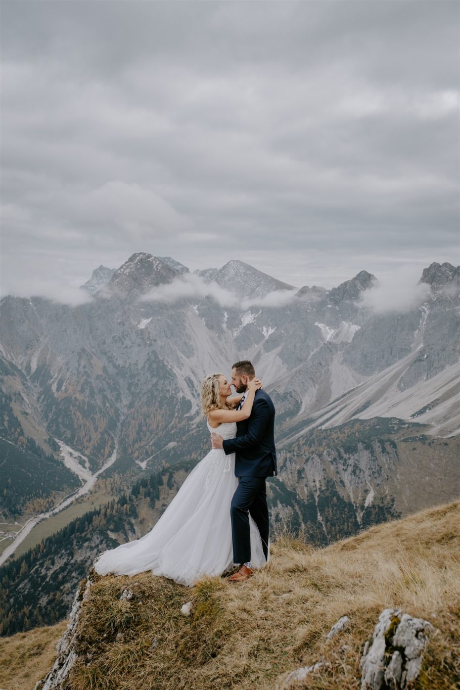 Bride and groom kiss on a mountain top with the Tyrol mountains in the distance behind them