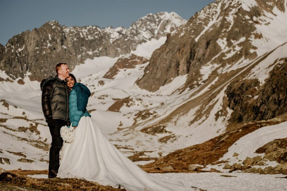 High alpine views to resemble Chamonix, bride and groom stand in the snow in down jackets over their wedding clothes