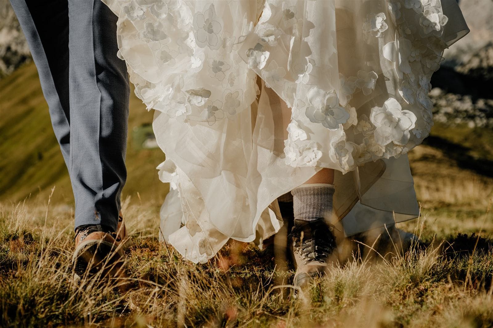 Close up of the feet of a bride and groom wearing hiking boots and walking through long grass.