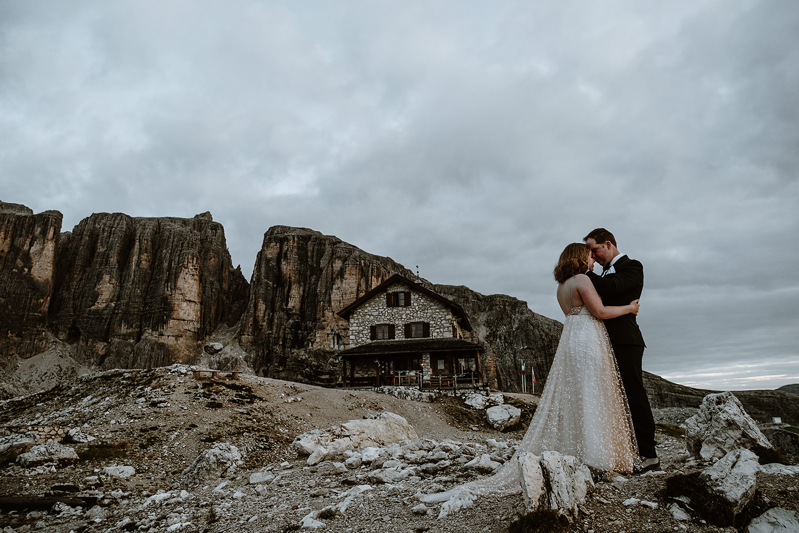 A mountain hut elopement location in the Dolomites. A bride and groom in a tight embrace with a stone mountain hut and tall mountains in the distance.