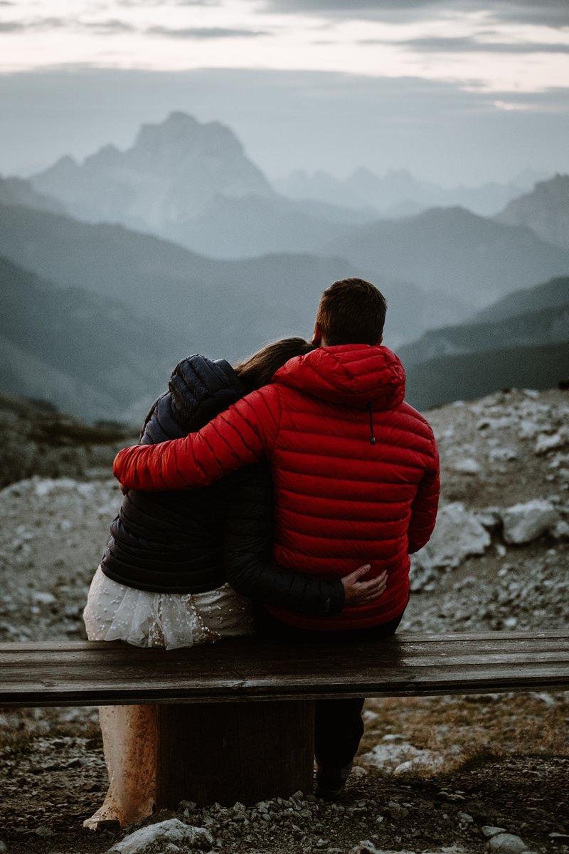 Bride and groom sitting on a bench in the Dolomite mountains in Italy wearing down jackets and looking out across the mountains