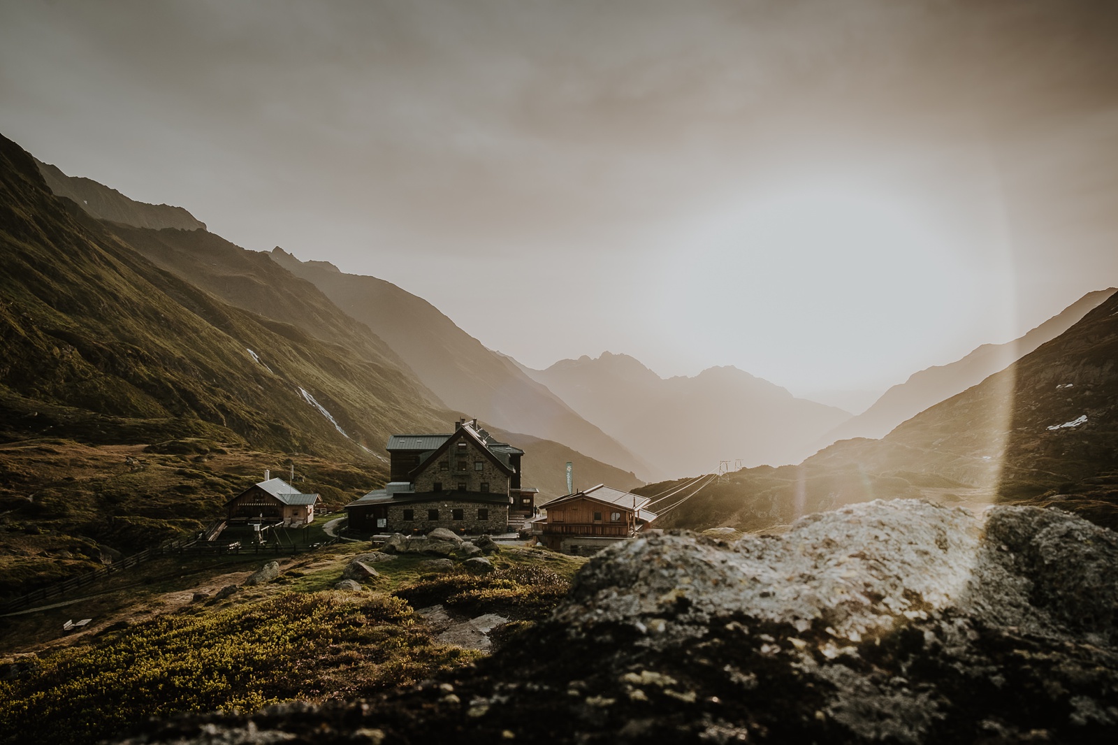 A mountain hut in the Austrian Alps at sunrise with green meadows and rocks in the foreground and high mountains in the distance.