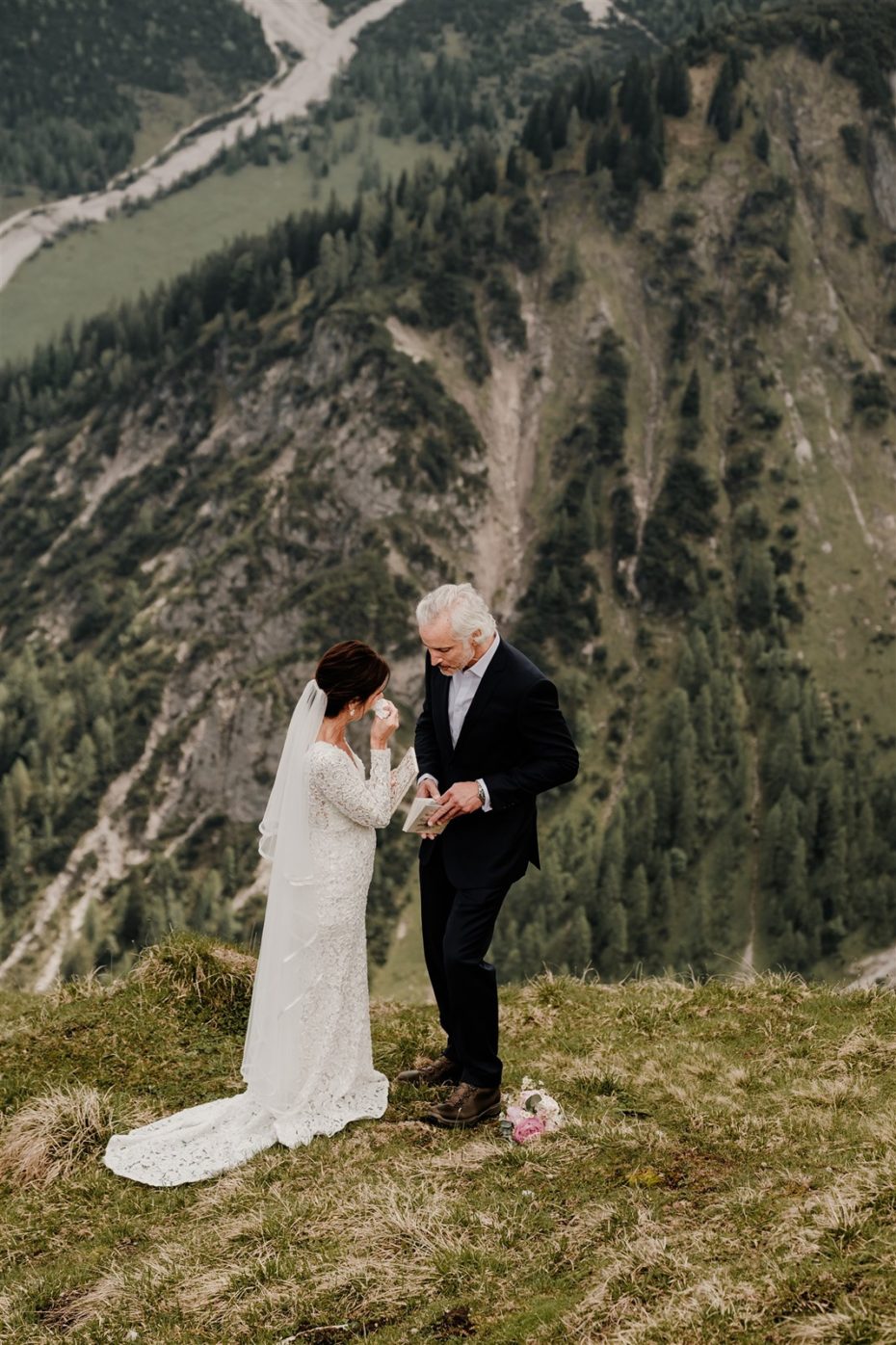 Bride wipes a tear from her face as groom reads wedding vows on top of a mountain in Seefeld