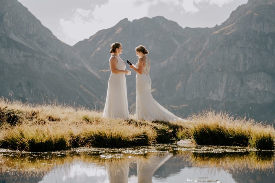 Two brides stand in an alpine meadow reading their wedding vows to one another