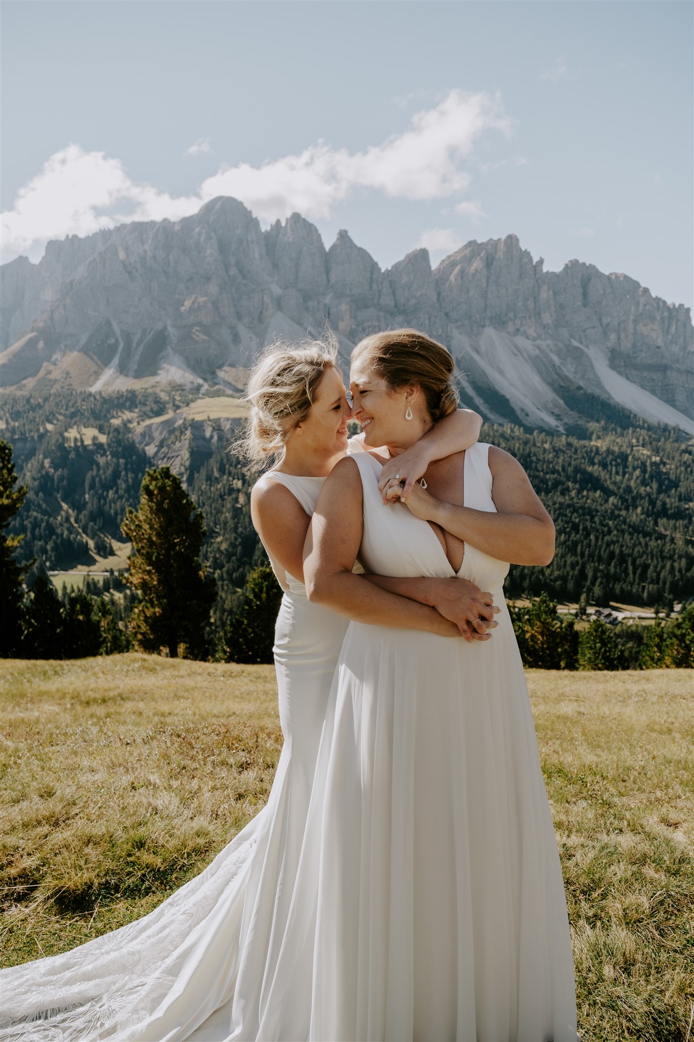 One bride stands behind her new wife, wrapping her arms around her waist, as the turn to each other and smile with the Dolomites in the background behind them.
