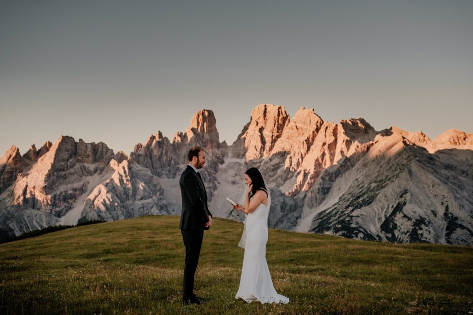 A bride and groom reading their vows to one another in a green meadow with the glowing Dolomite mountains behind them at sunrise