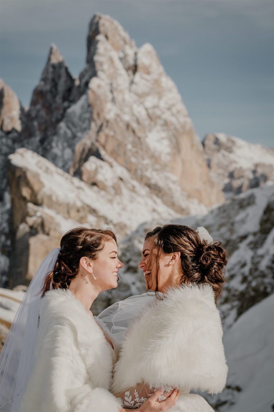 Two brides laugh and embrace in front of Seceda mountain in the Dolomites in winter