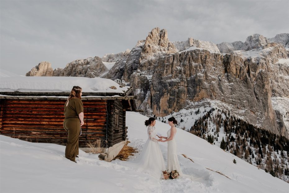 Two brides stand in the snow by a wooden mountain chalet in the Dolomites for their elopement ceremony