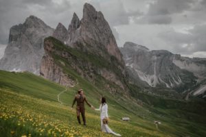 Groom leading bride up the steep mountain hillside of Seceda in the Dolomites with rocky knife-edge peaks in the distance