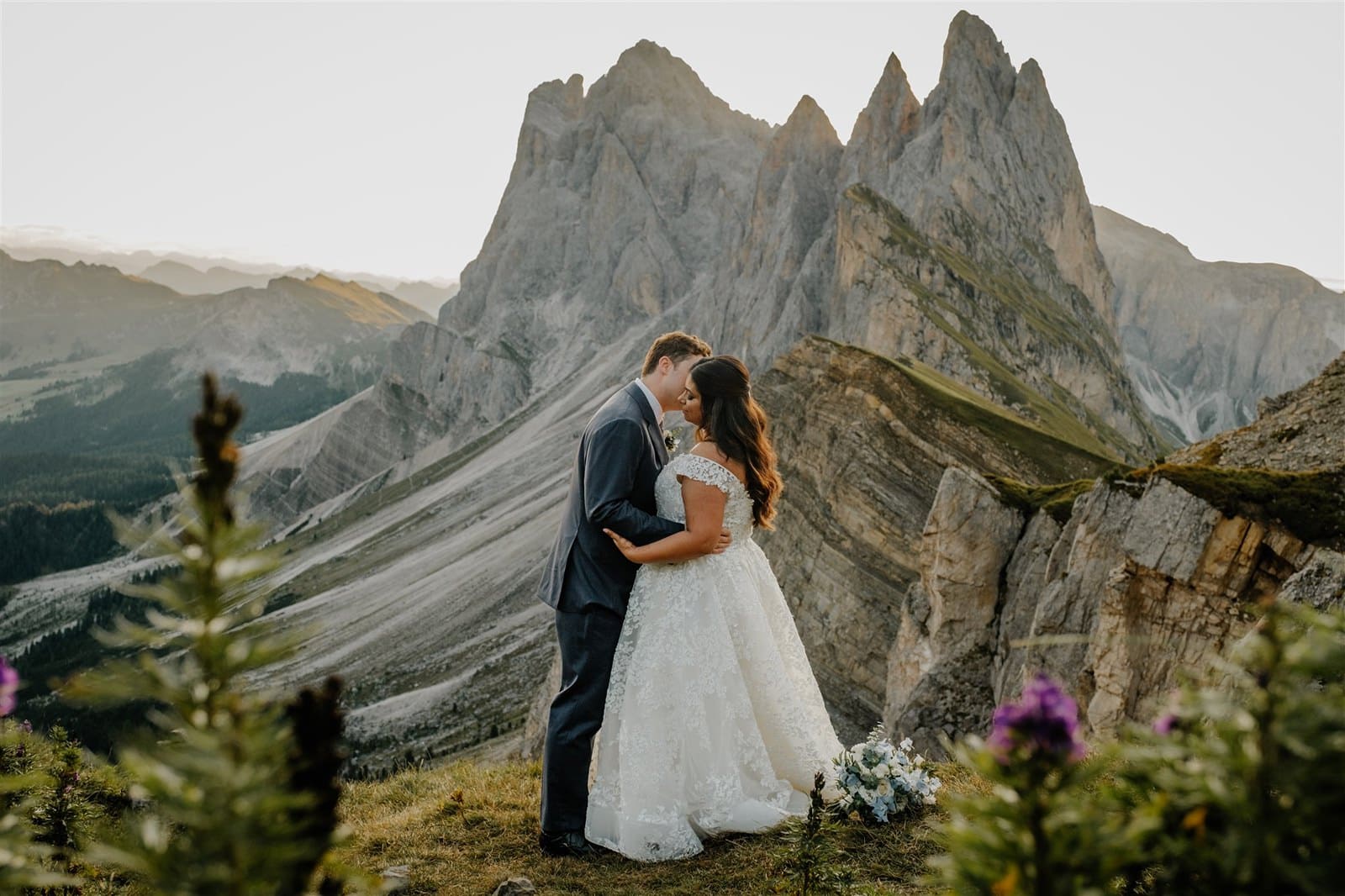 Bride and groom standing in a field of purple wildflowers kiss. Behind them is the famous Seceda mountain in the Dolomites.