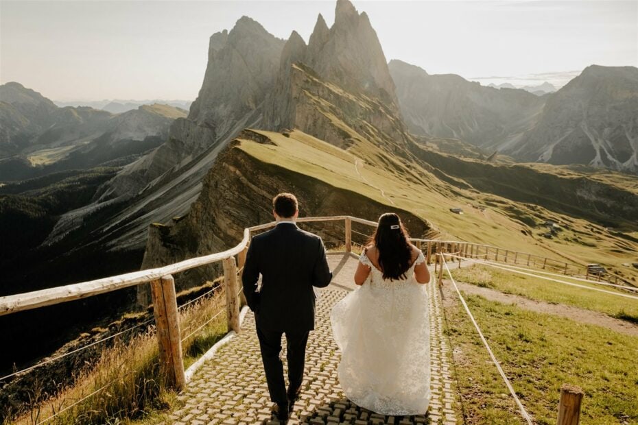 A photo showing a bride and groom walking downhill on a fenced footpath on the Seceda mountain in the Dolomites