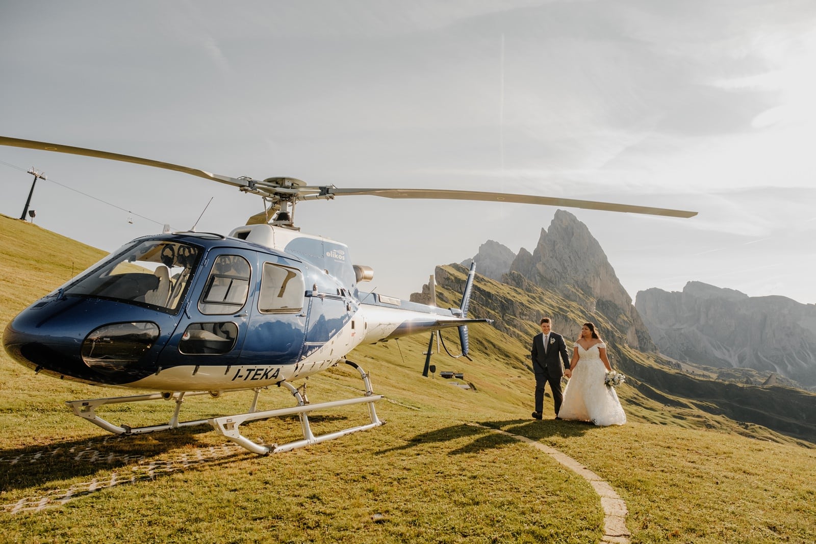 Bride and groom walk up to a blue helicopter that is parked on the hillside of the Seceda mountain in the Dolomites