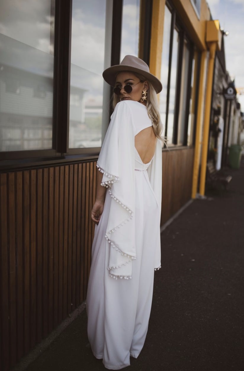 Bride wearing brown hat and while jumpsuit with cape-style sleeves with boho pom-pom details