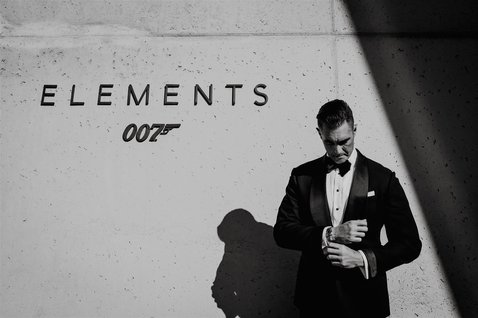 Groom in a black tuxedo standing by a concrete wall with 007 elements written on it