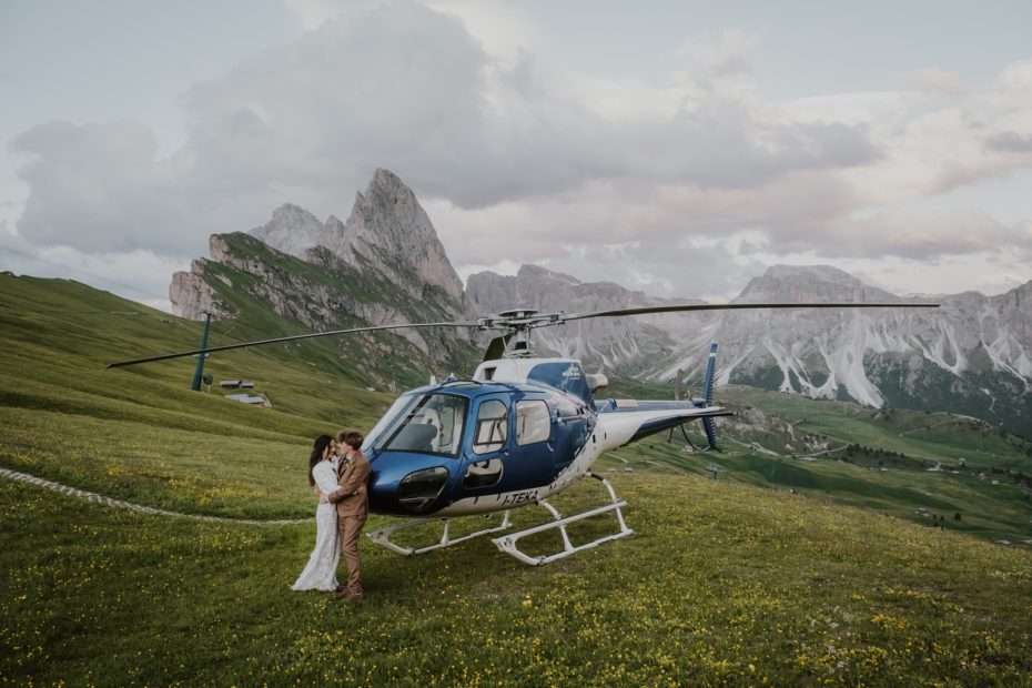 Bride and groom embracing while leaning on the nose of a helicopter on the edge of Seceda in the Italian Dolomites