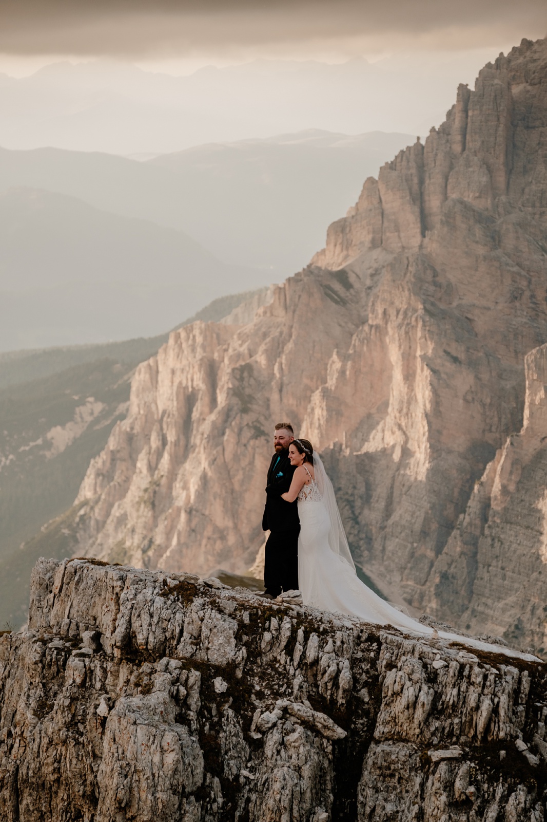 Bride and groom stand on the edge of a cliff and the bride embraces the groom from behind at sunset as the mountains glow pink behind them near a mountain refuge in the Dolomites.