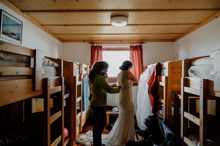 Women helps a bride button her wedding dress in a bunk room in a mountain hut in the Italian Dolomites