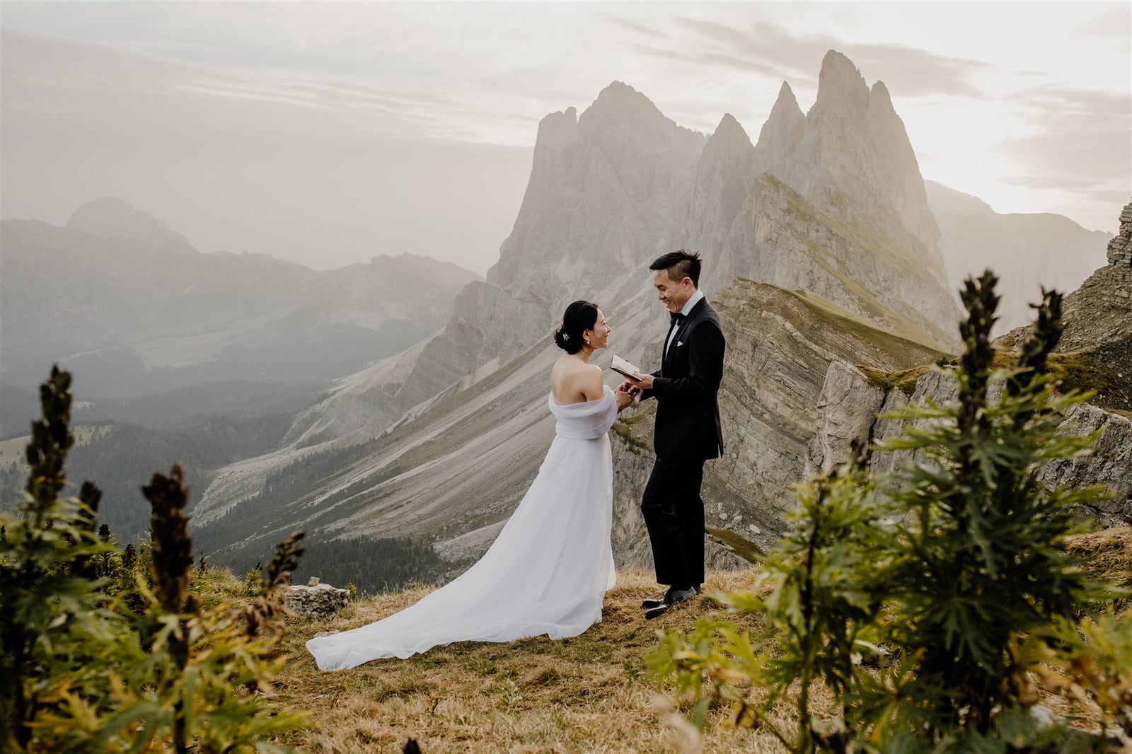 Asian bride and groom read wedding vows to one another standing in a meadow of wildflowers with the Seceda mountain in the distance behind them